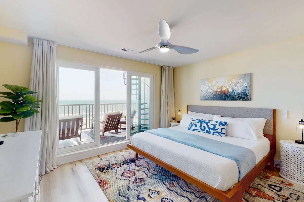 This image showcases a serene bedroom with a king bed adorned with blue accents, large windows that fill the space with natural light, and a balcony with seating that offers a glimpse of the ocean. A colorful rug adds warmth and personality, and the overall mood is tranquil and inviting.