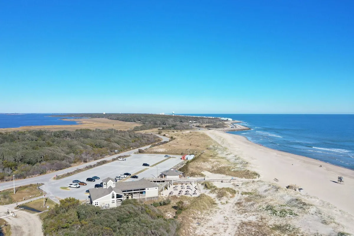 A coastal landscape featuring a beachfront restaurant, parking lot, and ocean, with early evening light hinting at upscale dining and social possibilities by the sea.