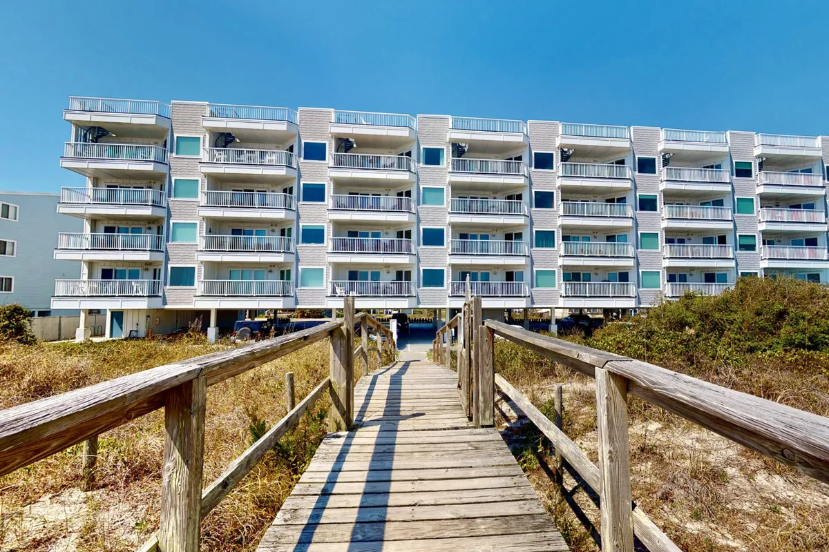 A welcoming exterior shot showing a wooden walkway leading to the building’s balconies with surrounding greenery. This perspective emphasizes easy beach access and outdoor relaxation areas, with the structure’s inviting balconies hinting at private retreats and community gatherings.