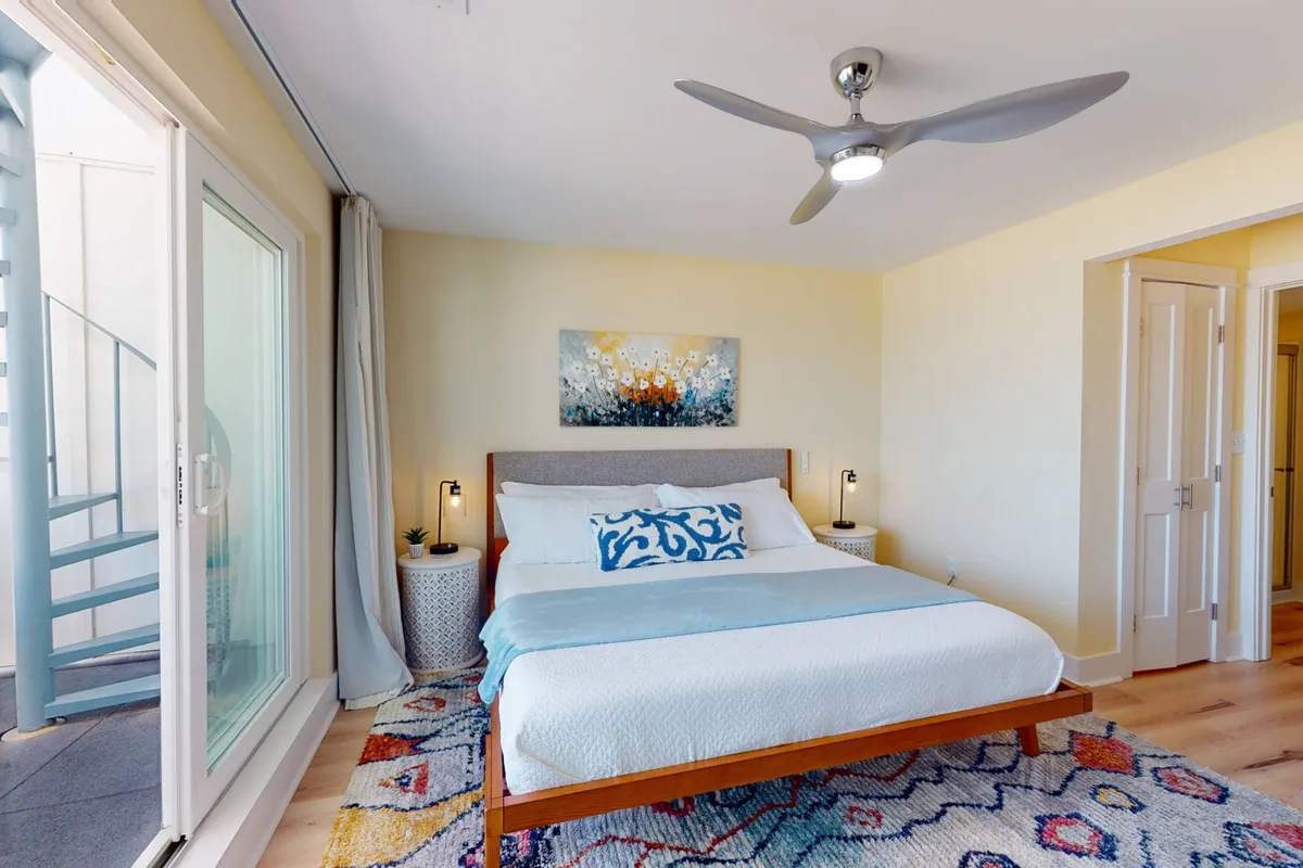 A cozy and serene bedroom featuring a bed with plush decorative pillows, an art piece above the headboard, a ceiling fan, and a colorful area rug. Natural light bathes the room, creating an inviting environment for relaxation and restful sleep.