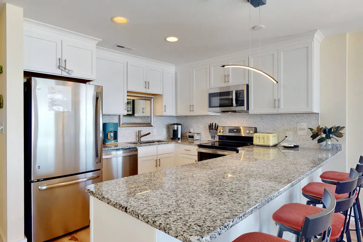 A modern kitchen with a granite countertop, stainless steel appliances, stylish bar stools, and a contemporary light fixture, suggesting both functionality and luxury for meal preparation and coffee breaks.