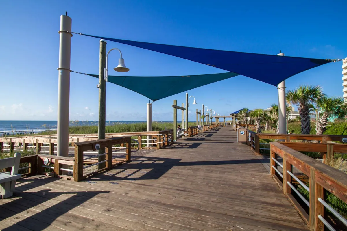 A scenic exterior showing the wooden boardwalk lined with colorful shade sails and surrounded by palm trees, all set against a view of the ocean. The inviting setting highlights the accessible pathways and iconic features of the area.