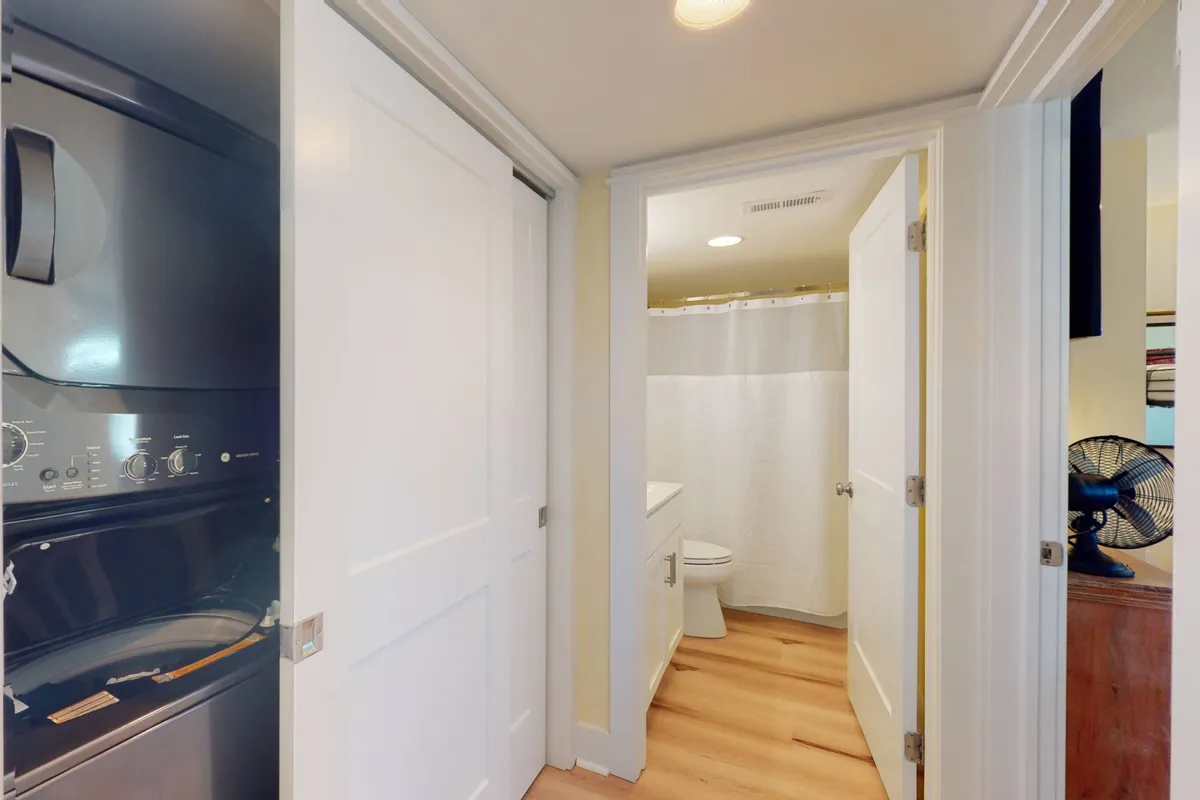 A bright and efficient laundry area featuring a large-capacity washer and dryer set against wooden flooring. The adjacent bathroom entrance and contemporary light fixtures complete the practical, modern vibe.