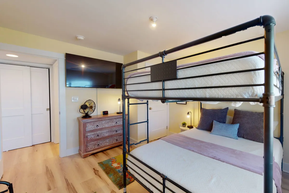 This room features a bunk bed setup with a queen-sized lower bunk and a single top bunk, complemented by a dresser, wall-mounted TV, and ceiling fan. The casual and family-friendly decor makes it perfect for children and families, and the space feels intimate and functional.