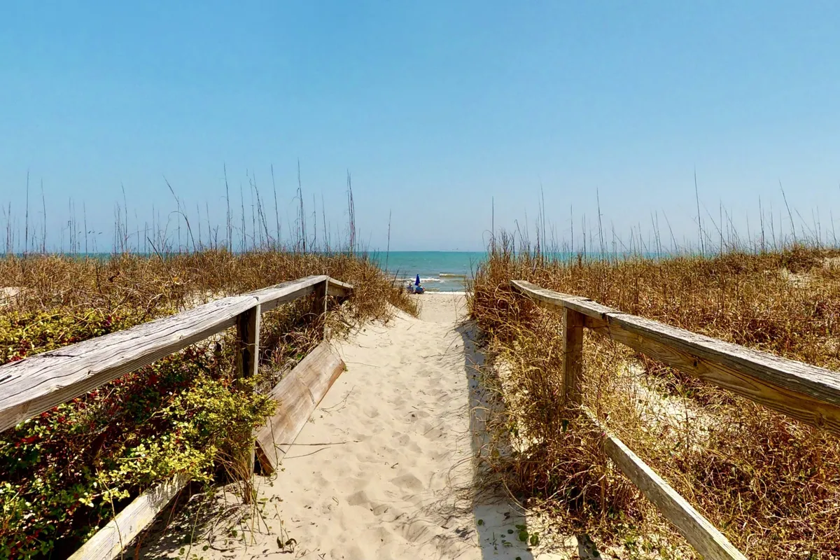 A wooden pathway leads through dune grass toward a serene beach and the ocean, bathed in gentle sunlight. The scene is peaceful, evoking a sense of romantic seclusion and connection to nature.