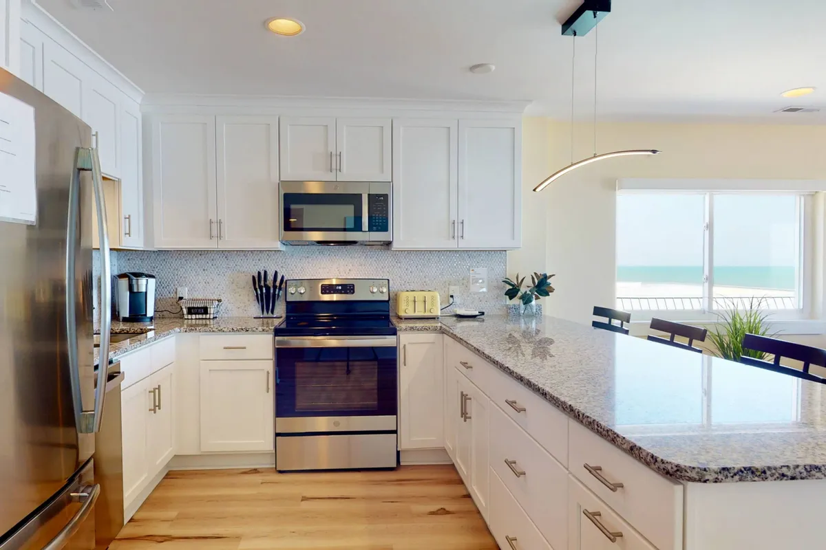 A sleek, modern kitchen showcasing granite countertops, stainless steel appliances, pendant lighting, and a large window with ocean views. Bar stools line the counter, inviting casual gatherings and meals.