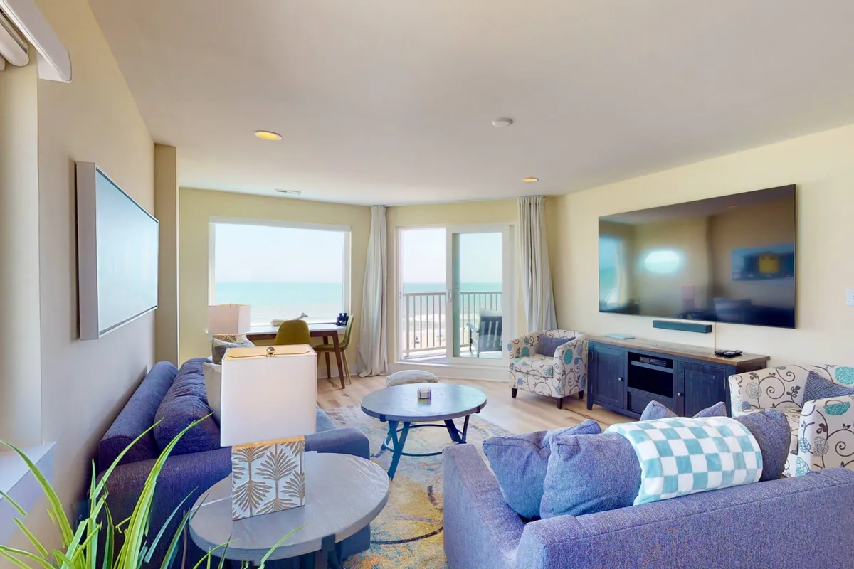 A sun-filled living room featuring a comfortable seating area oriented toward a large entertainment center, with expansive windows framing breathtaking ocean views. The space is open and airy, fostering a sense of togetherness and relaxation.