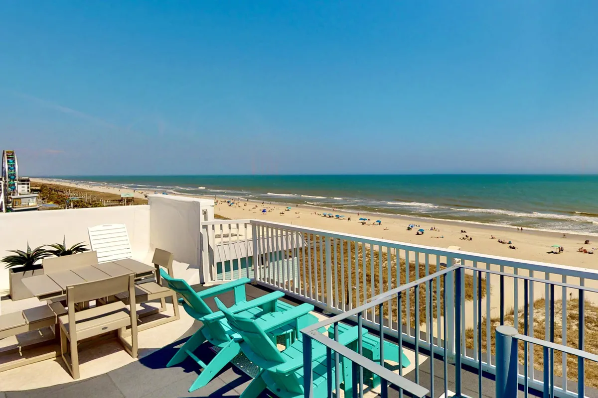 A serene oceanfront deck furnished with comfortable lounge chairs and a dining table, overlooking a sandy beach and the vast Atlantic Ocean. The soft glow of sunrise or sunset filters onto the deck, with the inviting ocean just steps away.