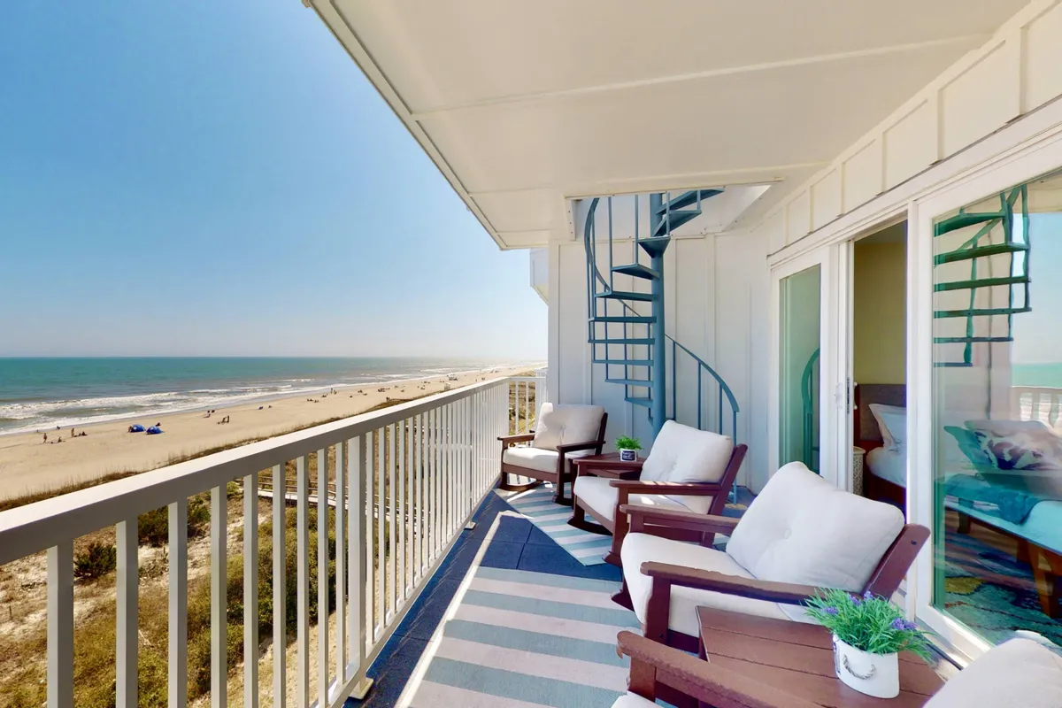 The hero image showcases the oceanfront balcony of a luxury top-floor condo at Carolina Beach. The scene highlights a spacious outdoor seating area, a striking spiral staircase leading to a private rooftop sundeck, and an expansive view of the sandy beach and sparkling blue Atlantic. The tranquil atmosphere invites guests to sunbathe, relax with a book, or simply soak in the unobstructed sunrise and ocean breezes.