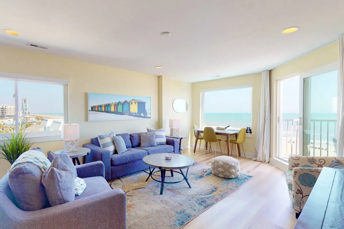 An inviting open-plan living and dining area, highlighted by a plush sofa, coffee table, tasteful coastal artwork, and a dining table adjoining the main living space. Large windows flood the room with natural light and ocean vistas.