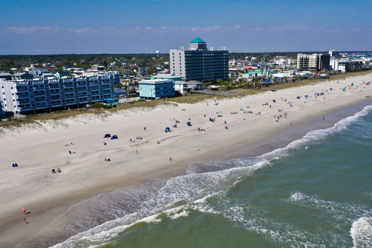 A vibrant exterior scene showing the beach with lively waves, people sunbathing, colorful umbrellas, and boardwalk buildings in the background. The atmosphere is bustling and joyful, suggesting plenty of family-friendly activities and easy beach access.